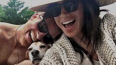 A selfie of Harry and Meghan laughing in sunglasses and cuddling a pet dog.