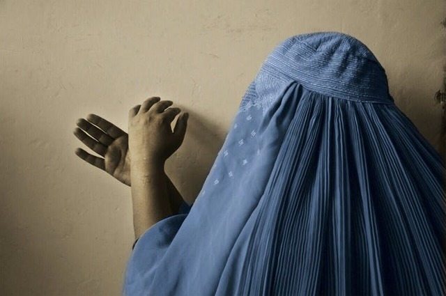 A woman in full burqa, anonymised, makes hand signs against a blank wall.